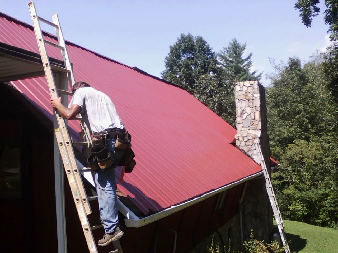 Roofer on a ladder beside a red metal roof and stone chimney — Soffit & Fascia Repair by Roofing Brothers in Battle Lake